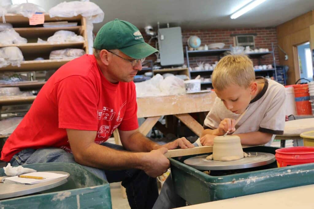 A photo of a teacher helping a young student refine a piece on a pottery wheel at the Clay Center in Greeley