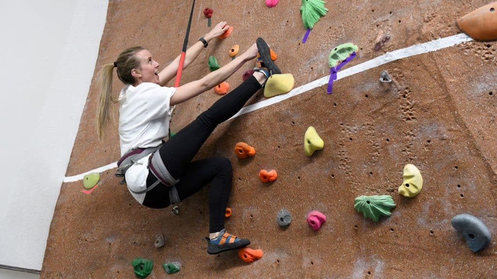 A young white woman with a pony tail, wearing a white t-shirt and black leggings, extends her leg to find a foothold on the colorful Greeley Rec Center climbing wall.