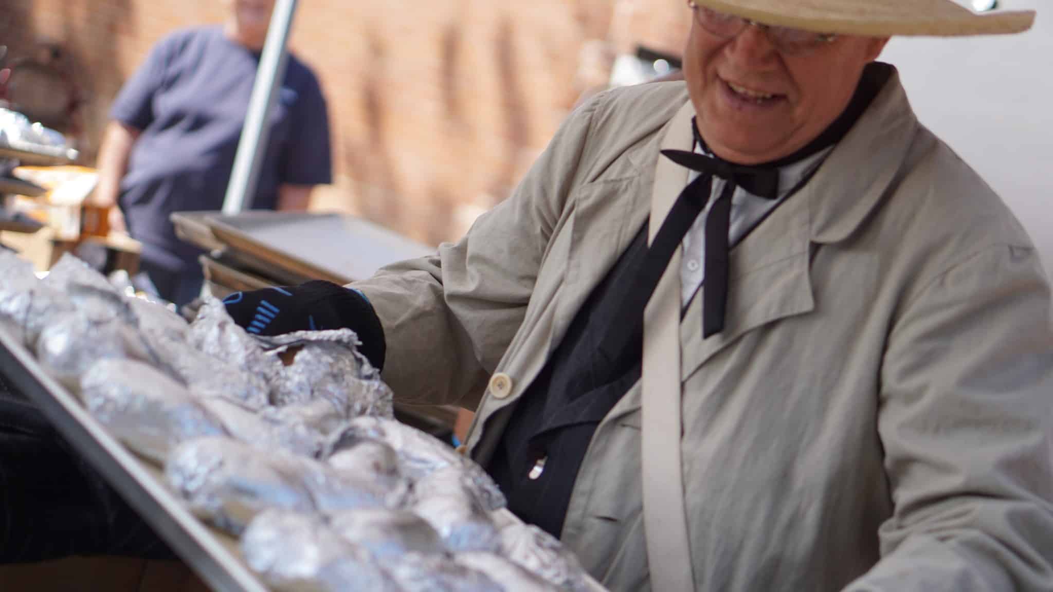 A man in a straw hat and homesteader costume lifts an extra large baking sheet covered in several dozen foil-wrapped baked potatoes