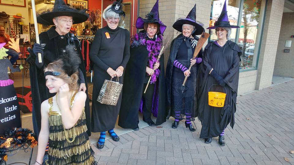 A young girl in a costume collects candy from a group of older women dressed as witches in black and purple at the Trick or Treat Street event in Greeley, Colorado