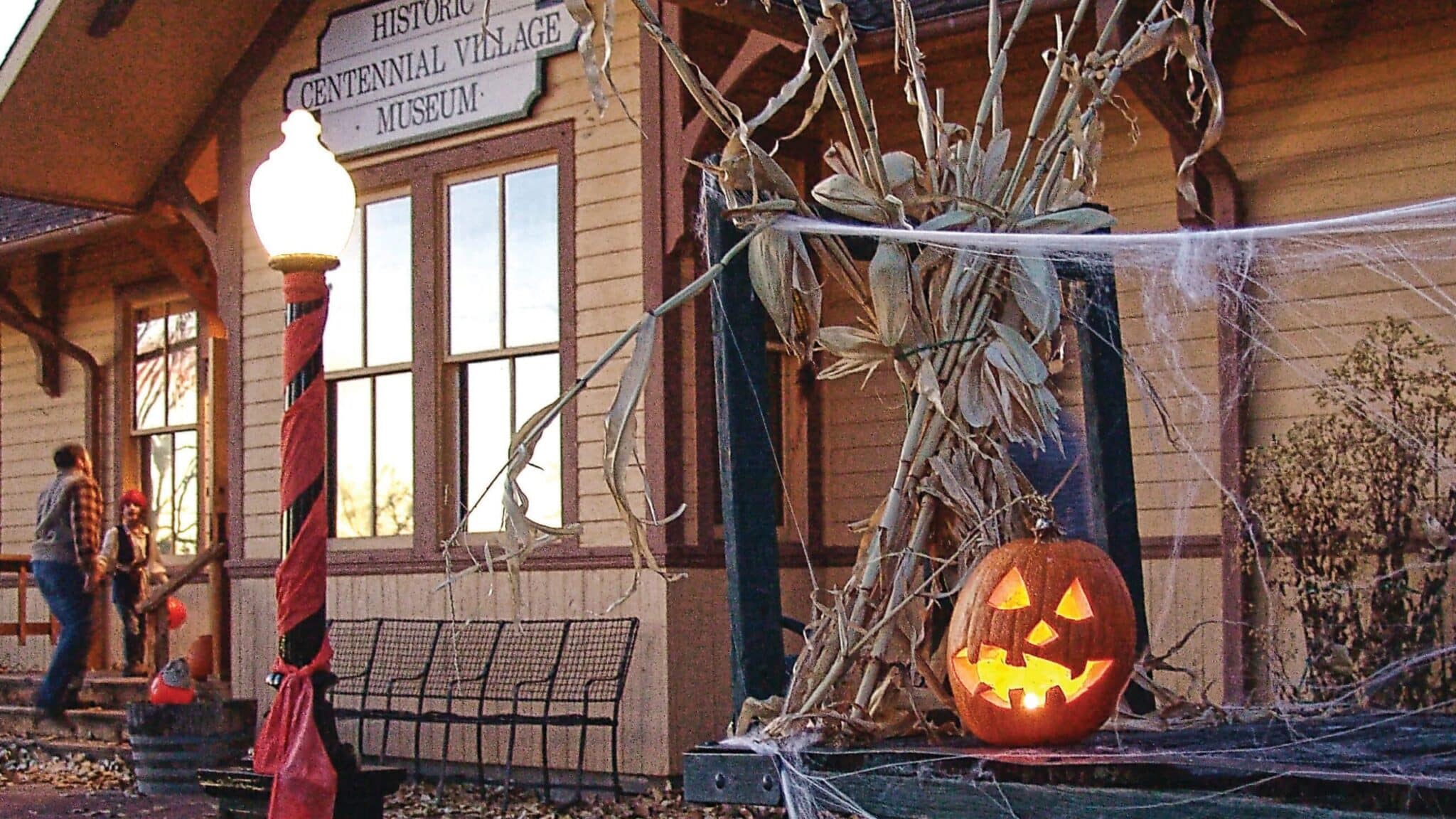 An outdoor photo of the entrance to the Historic Centennial Village Museum in Greeley, Colorado. The museum is decorated for fall with cornstalks, cobwebs, and a lit jack-o-lanturn,