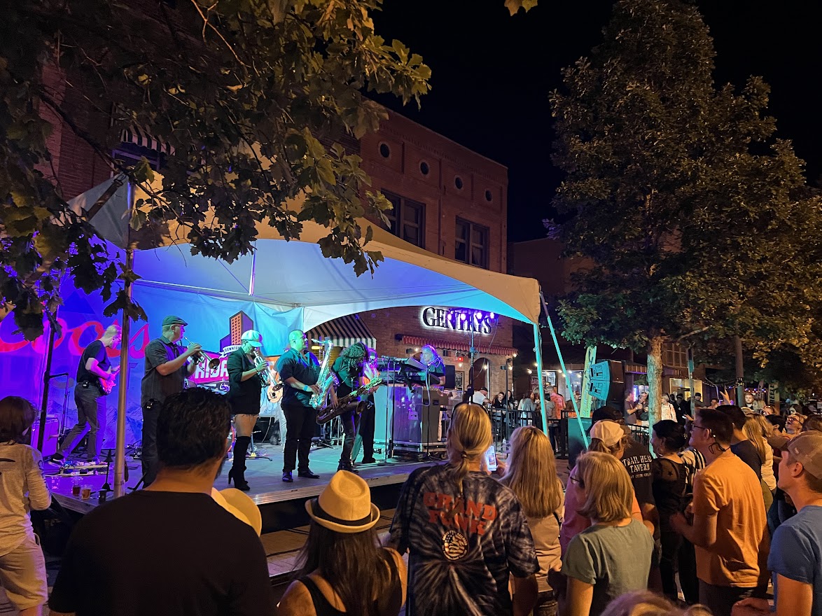 A large crowd stands outside in the dark in downtown Greeley, Colorado, as a large band plays on a stage lit in purple and green lights