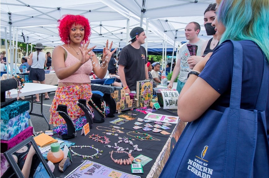 A young black woman with curly fuchsia colored hair chats with UNC student customers across a vendor table covered in jewelry