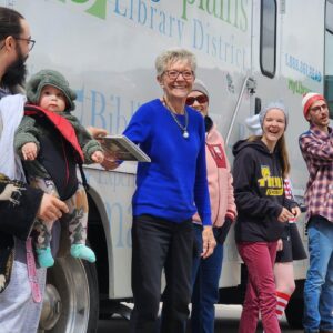 An all-ages group of Greeley community volunteers stand in front of a High Plains Library District bus, passing books from hand to hand.